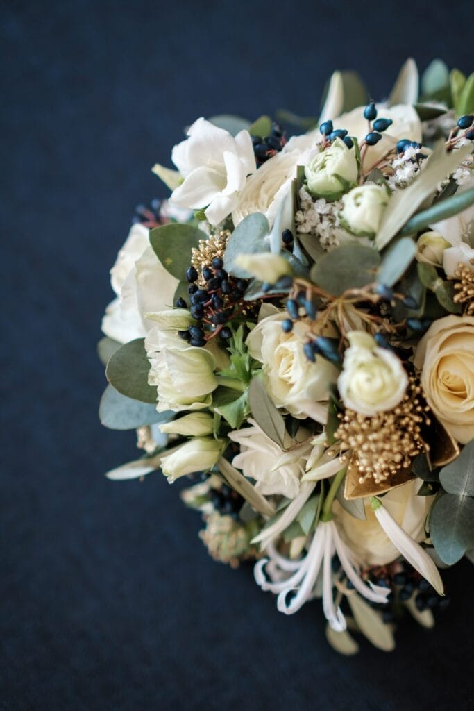 Hochzeitskrone Ein Strauß weißer Rosen, grüner Blätter und kleiner dunkler Beeren vor dunklem Hintergrund fängt bezaubernde Momente ein, die an natürliche Hochzeitsfotos erinnern.