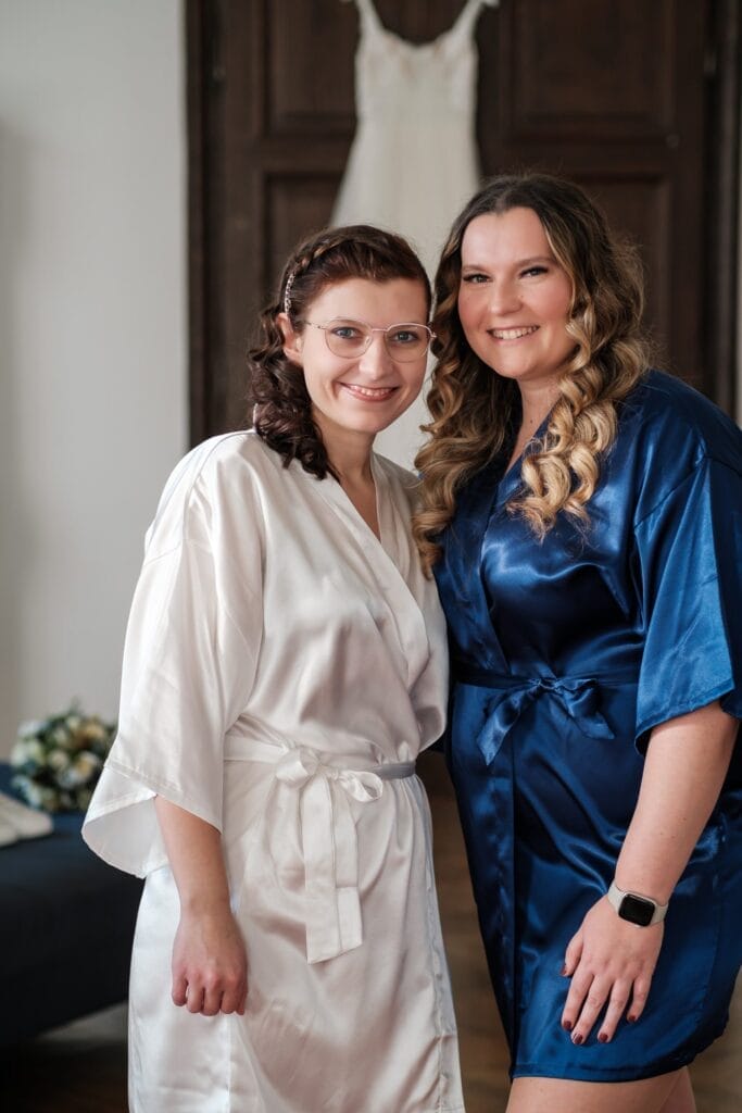 Hochzeitskrone Zwei lächelnde Frauen in Satinroben, eine weiß und eine blau, teilen einen bezaubernden Moment im Innenbereich. Im Hintergrund sind ein weißes Kleid und ein Blumenstrauß zu sehen, die die Essenz natürlicher Hochzeitsfotos perfekt einfangen.