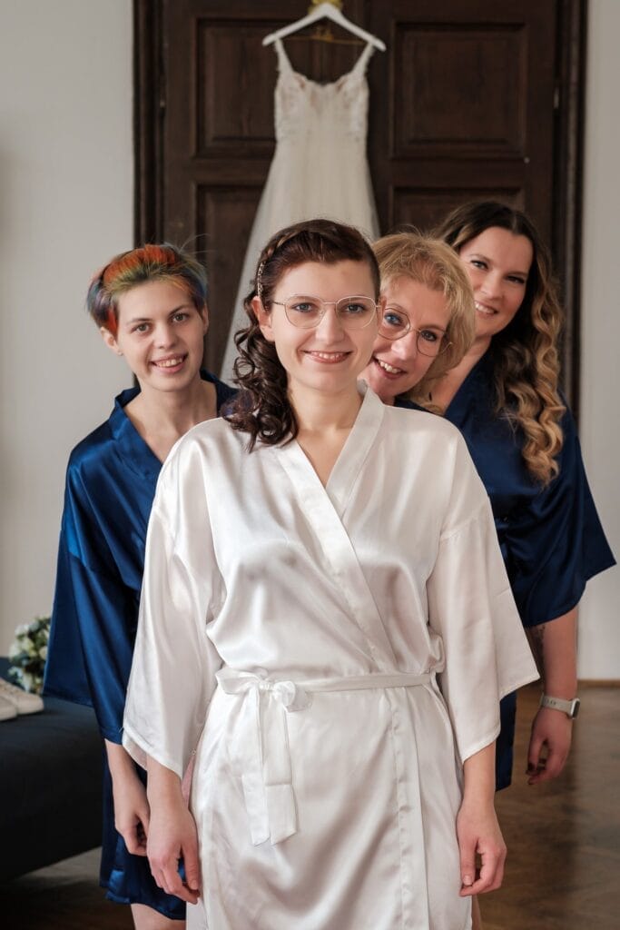 Hochzeitskrone Vier Frauen in Roben stehen lächelnd in einer Reihe im Innenbereich, während hinter ihnen ein Brautkleid an einer Tür hängt. Diese Szene fängt bezaubernde Momente der Hochzeitsfotografie in Niederösterreich ein, wo natürliche Schönheit und Freude mühelos gefeiert werden.