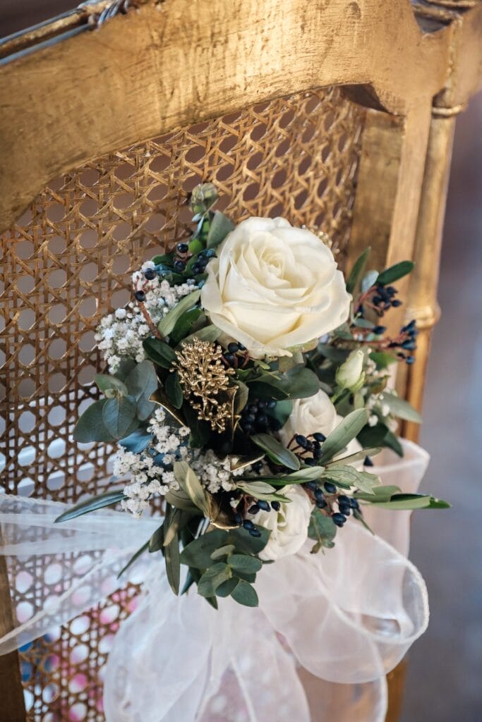 Hochzeitskrone Weiße Rosen, Schleierkraut und Grünpflanzen, elegant arrangiert auf einem goldverzierten Stuhl mit einer hauchdünnen weißen Schleife, fangen bezaubernde Momente in dieser exquisiten Umgebung ein.