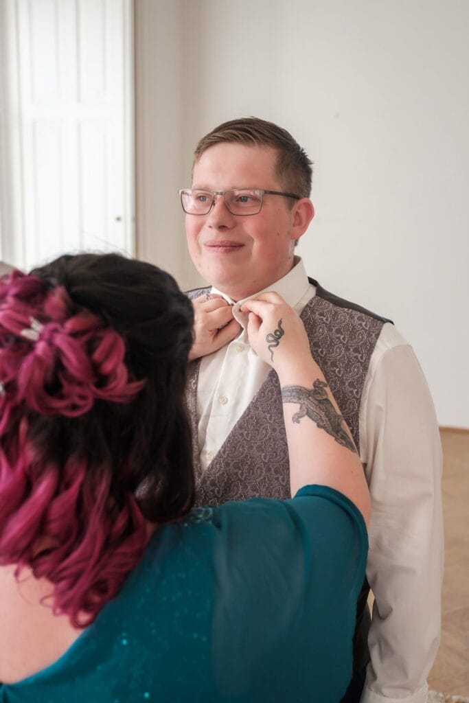 Hochzeitskrone Eine Person mit rosa Haaren hilft einer bebrillten Person in formeller Kleidung dabei, die Krawatte zurechtzurücken, und fängt damit einen dieser bezaubernden Momente ein, die man in der Hochzeitsfotografie in Niederösterreich oft sieht.