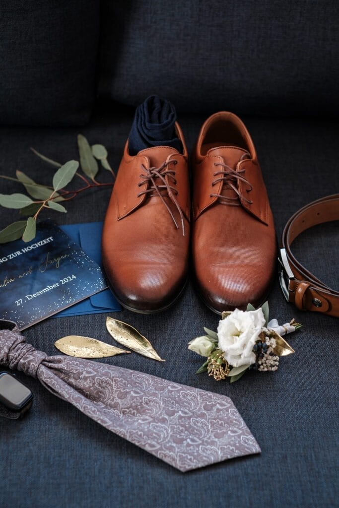 Hochzeitskrone Braune Lederschuhe mit passendem Gürtel, gemusterter Krawatte und Boutonniere fangen bezaubernde Momente ein. Einladungen und dekorative Blätter sind kunstvoll auf einem dunklen Untergrund arrangiert und veranschaulichen so natürliche Hochzeitsfotos in Niederösterreich.