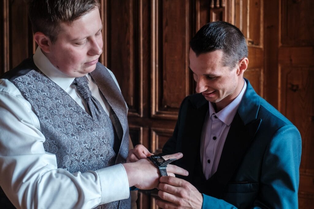 Hochzeitskrone Zwei Männer in formeller Kleidung, einer hilft dem anderen, eine Uhr an seinem Handgelenk zu befestigen. Sie stehen vor einer Holzvertäfelung und fangen mit konzentrierten Ausdrücken bezaubernde Momente ein – perfekt für natürliche Hochzeitsfotos in Niederösterreich.