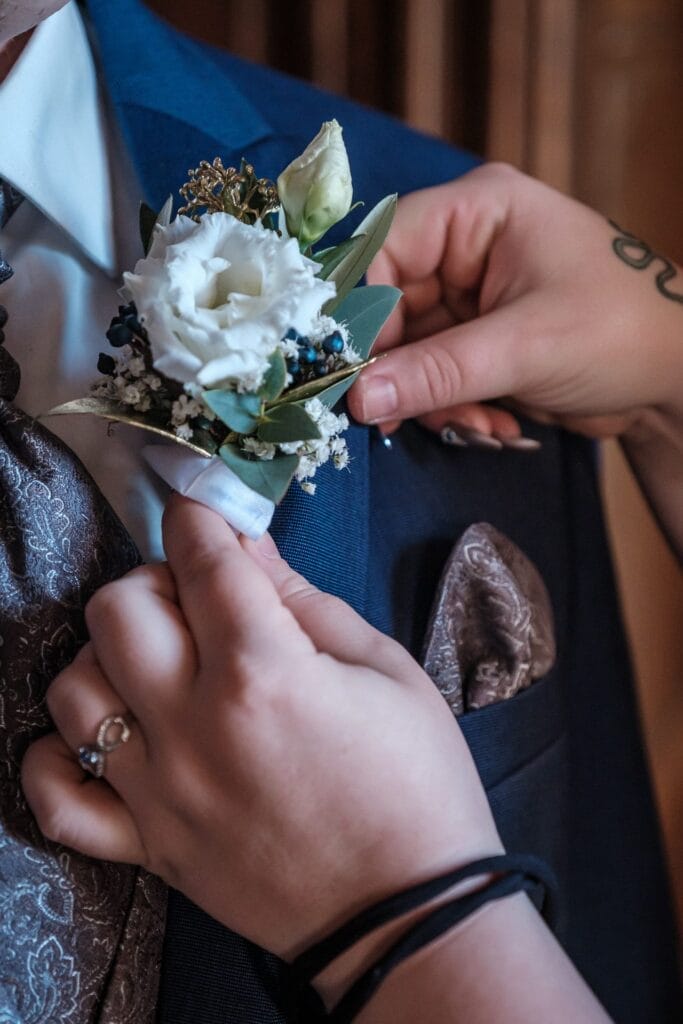 Hochzeitskrone Hände richten sorgfältig eine weiße Blumenansteckblume an einer Person in einem blauen Anzug und einer gemusterten Krawatte aus und fangen damit einen der bezaubernden Momente der Hochzeitsfotografie in Niederösterreich ein.