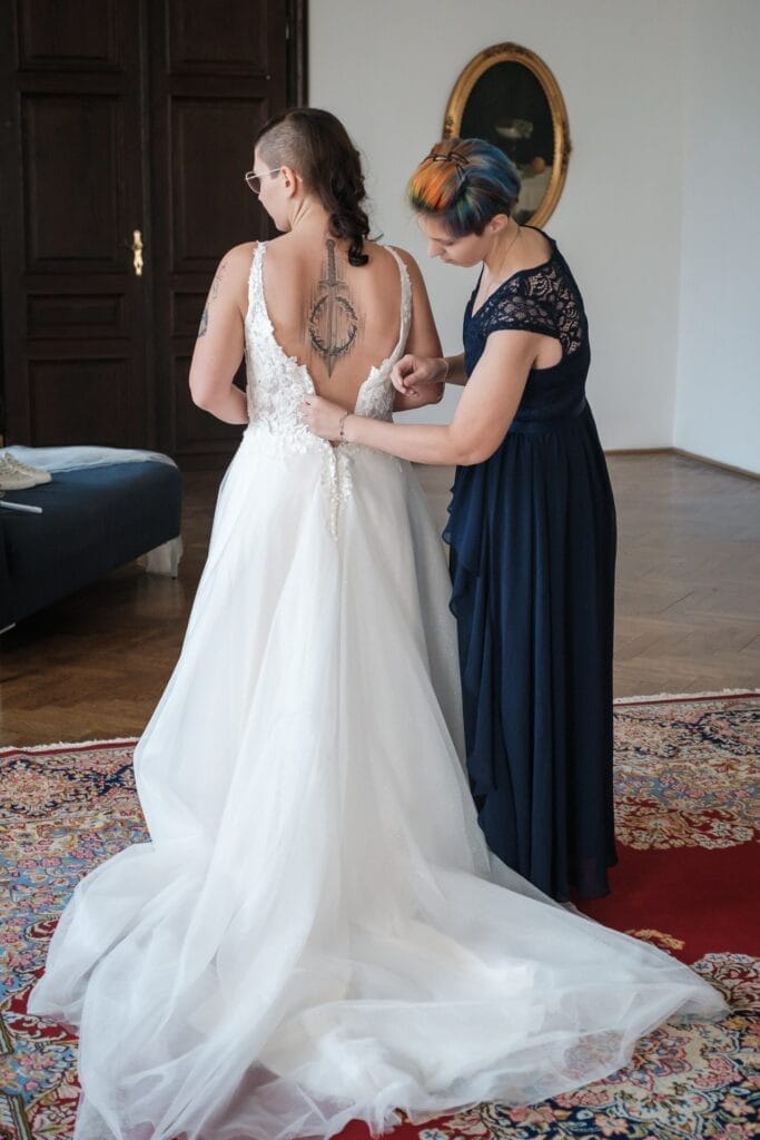 Hochzeitskrone Eine Frau im Hochzeitskleid lässt sich von einer anderen Frau im dunklen Kleid das Kleid zuknöpfen und fängt bezaubernde Momente auf einem gemusterten Teppich in einem Raum mit Holztüren und einem gerahmten Bild ein und ist ein Beispiel für natürliche Hochzeitsfotos.