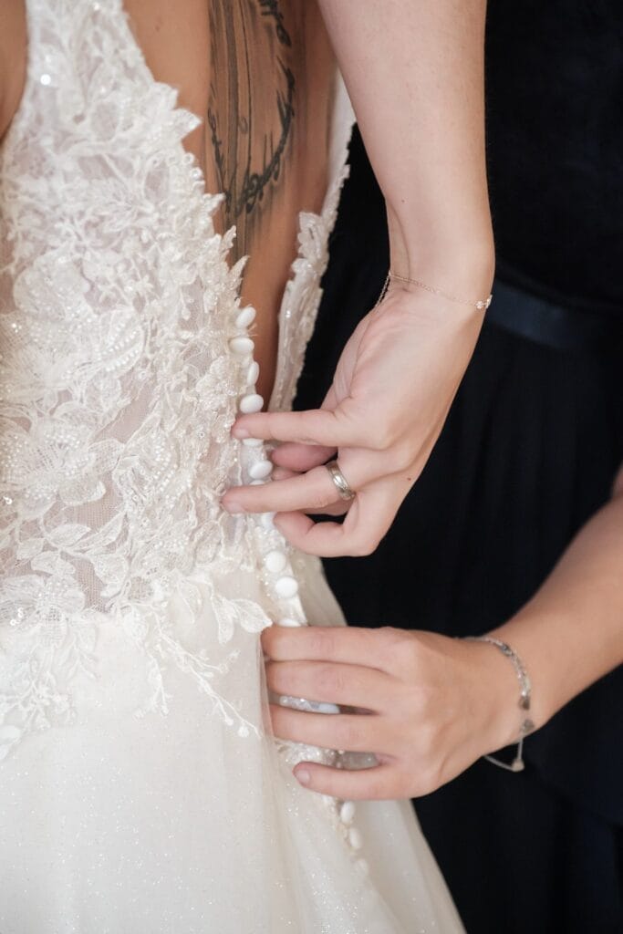 Hochzeitskrone Eine Person schließt die Knöpfe auf der Rückseite eines Brautkleides und gibt den Blick auf Spitzendetails und ein Tattoo auf dem Rücken der Trägerin frei – bezaubernde Momente, perfekt eingefangen von Hochzeitsfotografie in Niederösterreich.