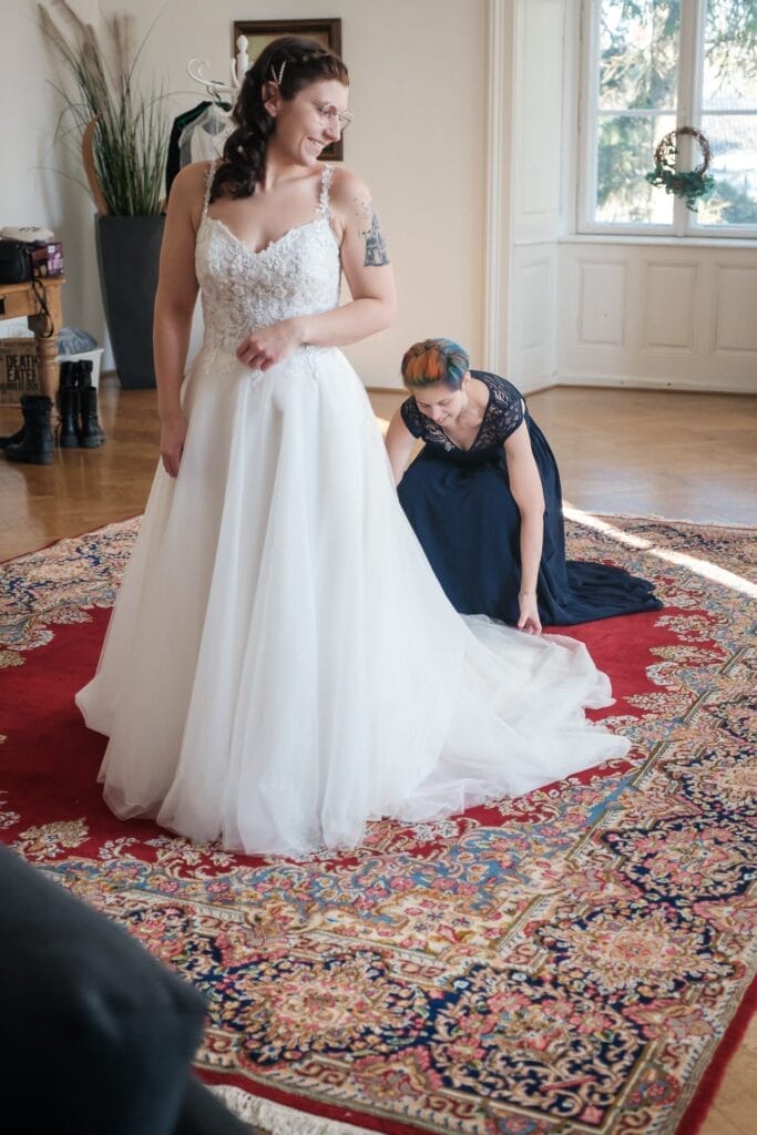 Hochzeitskrone In einem Raum mit großen Fenstern steht eine Braut in einem weißen Kleid auf einem rot gemusterten Teppich, während eine andere Person die Schleppe des Kleides zurechtrückt und so einen jener bezaubernden Momente einfängt, die typisch für natürliche Hochzeitsfotos sind.