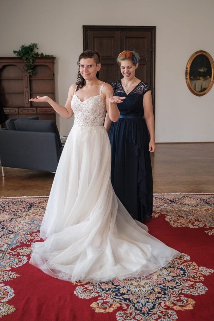 Hochzeitskrone In einem mit Holzmöbeln ausgestatteten Raum erleben zwei Frauen bezaubernde Momente. Die eine im Brautkleid zuckt verspielt mit den Schultern, während die andere im dunklen Kleid lächelnd hinter ihr steht. Sie liegen anmutig auf einem roten, gemusterten Teppich – perfekte natürliche Hochzeitsfotos aus Niederösterreich.