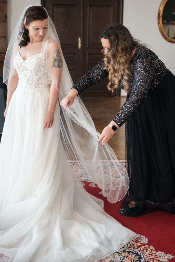 Hochzeitskrone In einer bezaubernden Szene von Hochzeitsfotografie in Niederösterreich steht die Braut in einem weißen Kleid, ihr Tattoo ist dezent sichtbar, auf einem roten Teppich. In der Nähe rückt eine Frau in einem schwarzen Kleid zwischen Holztüren zärtlich ihren Schleier zurecht und fängt bezaubernde Momente ein.