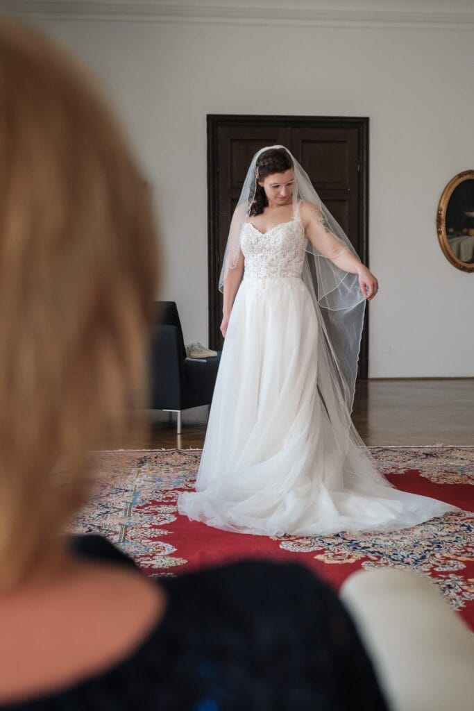 Hochzeitskrone Eine Braut in einem weißen Brautkleid und Schleier steht auf einem gemusterten Teppich und richtet ihr Kleid, während im Vordergrund eine andere Person unscharf zu sehen ist – so fängt diese bezaubernden natürlichen Hochzeitsfotos ein.