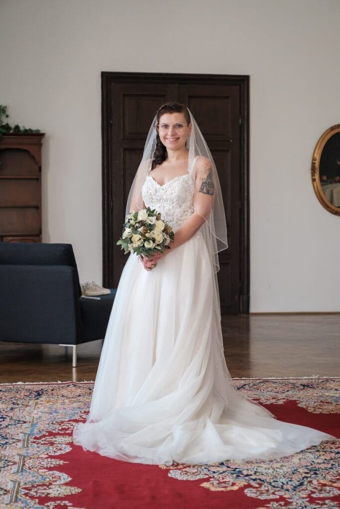 Hochzeitskrone Die Braut, in einem weißen Kleid und mit einem Blumenstrauß in der Hand, steht auf einem roten Teppich in einem Raum mit einer dunklen Tür und einem blauen Sofa und fängt bezaubernde Momente ein, die sich perfekt für natürliche Hochzeitsfotos eignen.