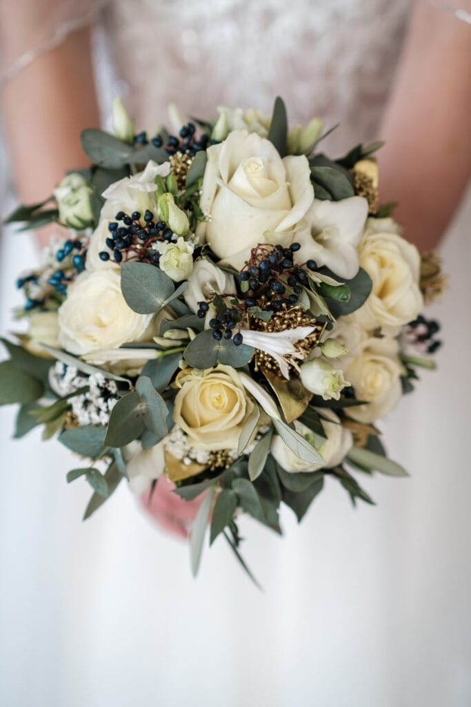 Hochzeitskrone Eine Person hält einen Brautstrauß mit weißen Rosen, Eukalyptusblättern und kleinen dunklen Beeren und fängt bezaubernde Momente einer Hochzeit ein.