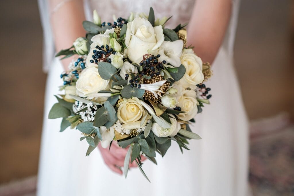 Hochzeitskrone Die Braut hält einen Strauß mit weißen Rosen, Grünpflanzen und kleinen dunklen Beeren in der Hand und trägt ein weißes Hochzeitskleid – sie fängt bezaubernde Momente ein durch natürliche Hochzeitsfotos in Niederösterreich.