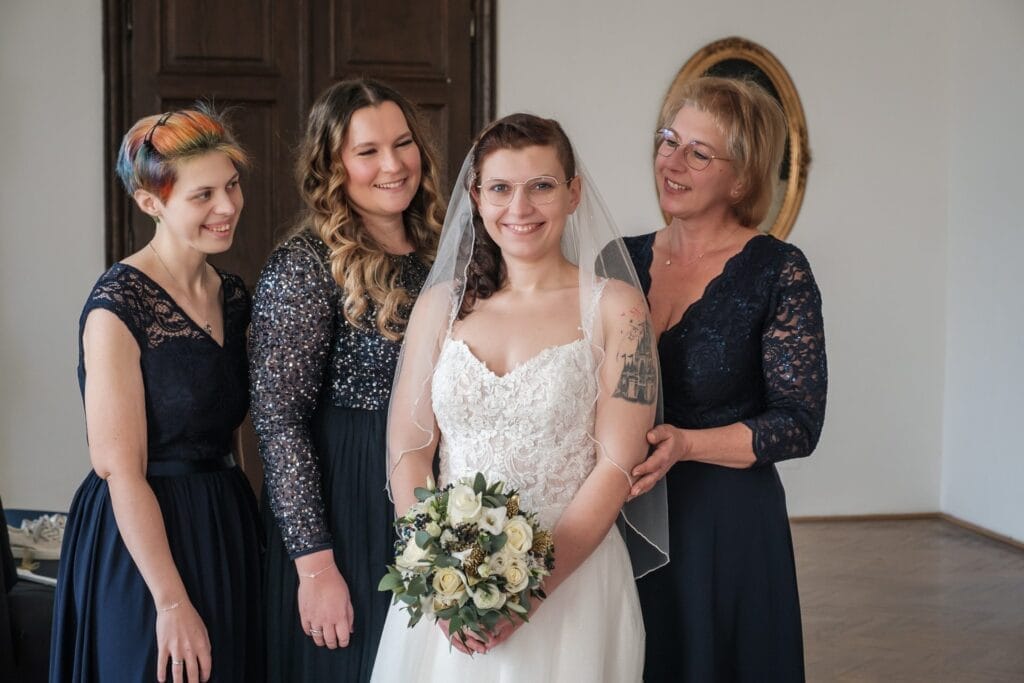 Hochzeitskrone Eine Braut in einem weißen Kleid mit einem Blumenstrauß steht mit drei Frauen in dunklen Kleidern zusammen, die alle lächeln und bezaubernde Momente einfangen. Sie befinden sich in einem Raum mit einer Holztür und einem Wandspiegel, perfekt für natürliche Hochzeitsfotos, die den Charme der Hochzeitsfotografie in Niederösterreich widerspiegeln.