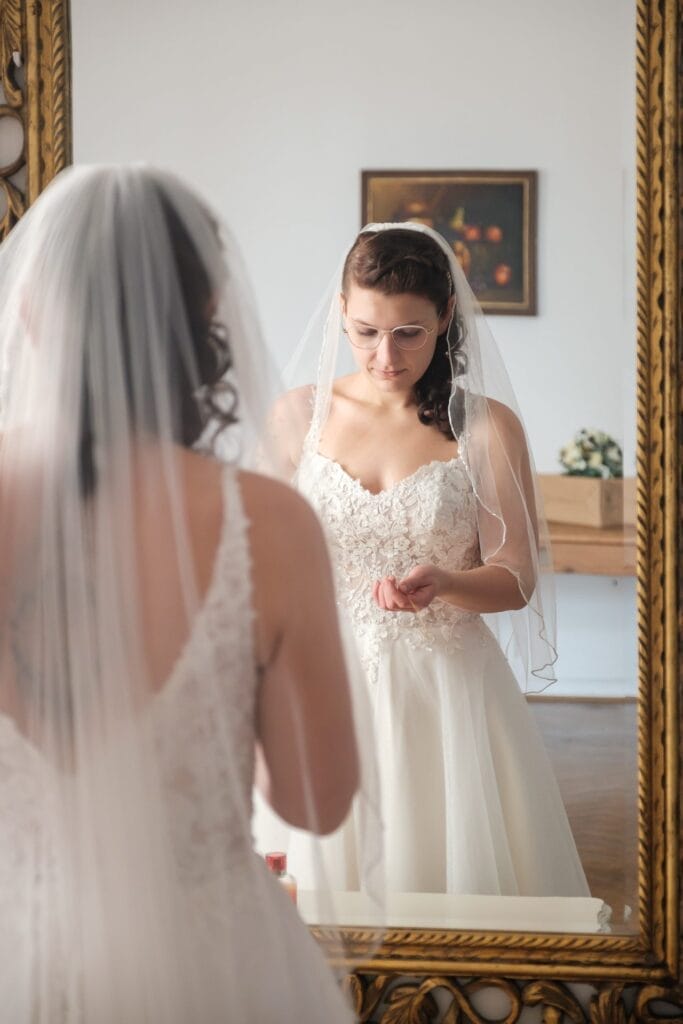 Hochzeitskrone Eine Braut in einem weißen Kleid und Schleier steht vor einem Spiegel, blickt nach unten und hält dabei einen kleinen Gegenstand in der Hand, und fängt einen dieser bezaubernden Momente ein, die sich perfekt für natürliche Hochzeitsfotos eignen.