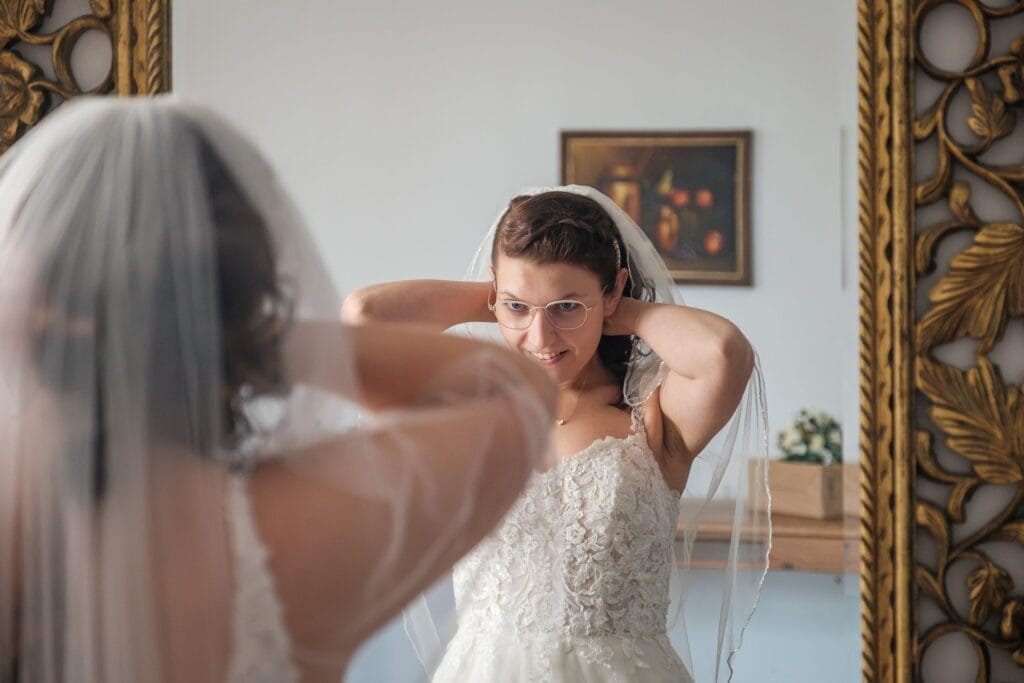 Hochzeitskrone Die Braut richtet ihren Schleier und lächelt dabei ihr Spiegelbild in einem dekorativen Spiegel an und fängt bezaubernde Momente purer Freude ein – perfekt für natürliche Hochzeitsfotos.