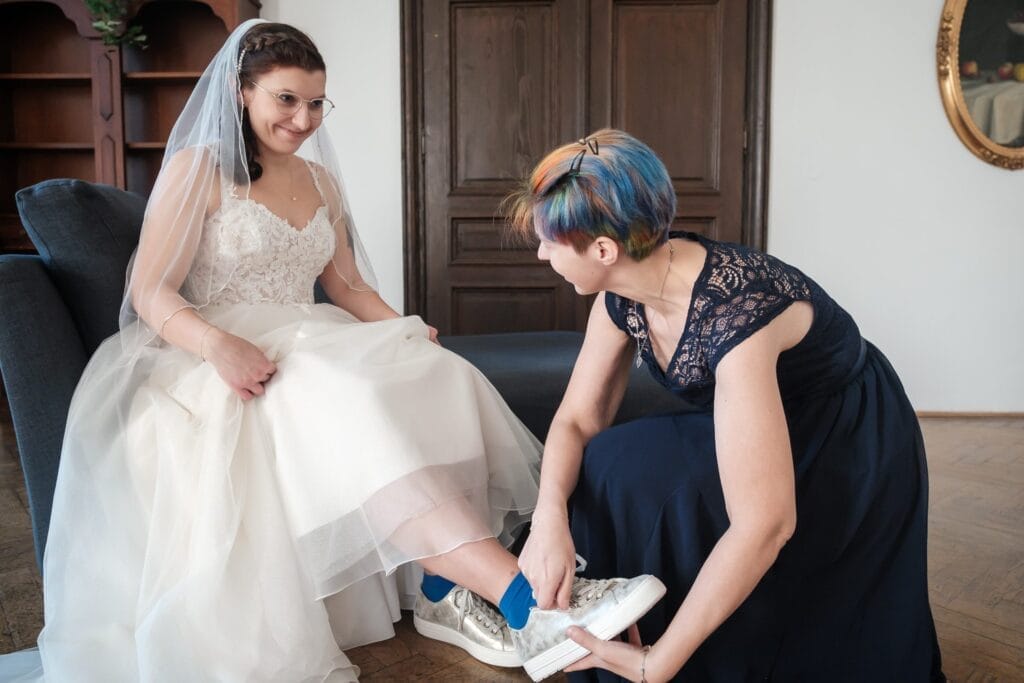 Hochzeitskrone Eine Braut in einem weißen Kleid und Schleier sitzt auf einem Sofa, während eine Frau ihre Schnürsenkel bindet und bezaubernde Momente festhält. Die glänzenden Turnschuhe der Braut mit blauen Socken verleihen einen Hauch von Charme und illustrieren perfekt natürliche Hochzeitsfotos für diese wertvollen Erinnerungen.