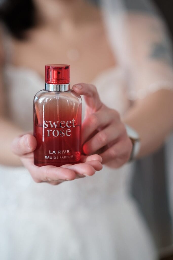 Hochzeitskrone Eine Person hält mit beiden Händen eine Flasche „Sweet Rose La Rive Eau de Parfum“ und fängt bezaubernde Momente ein, als ob es sich um eine natürliche Hochzeitsfotografie-Sitzung handelt.