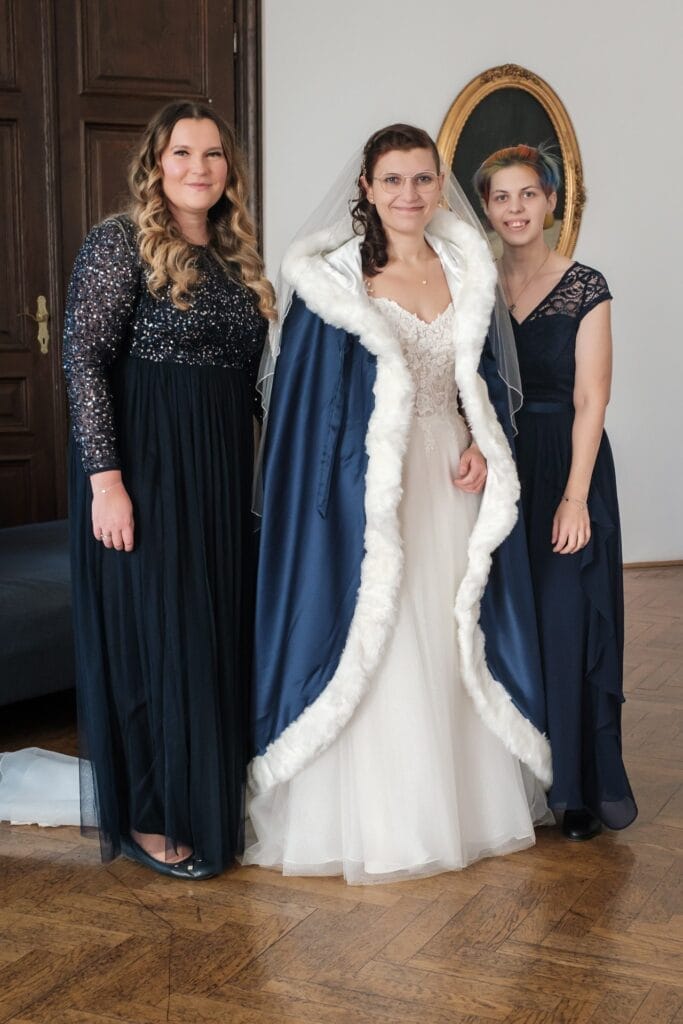 Hochzeitskrone Die Braut, in einem weißen Kleid und einem blauen Umhang, steht anmutig zwischen zwei Frauen in dunklen Kleidern und fängt bezaubernde Momente der Hochzeitsfotografie in Niederösterreich ein.