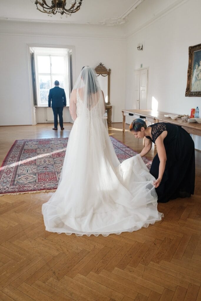 Hochzeitskrone Eine Braut in einem weißen Kleid und Schleier steht drinnen auf einem Teppich, während eine Frau ihr Kleid zurechtrückt. In der Nähe steht eine Person im Anzug an einem Fenster und fängt bezaubernde Momente natürlicher Hochzeitsfotos ein, die die Magie der Hochzeitsfotografie in Niederösterreich hervorheben.