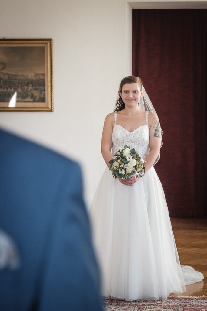 Hochzeitskrone Eine Braut in einem weißen Kleid mit einem Blumenstrauß steht drinnen und fängt bezaubernde Momente ein. Im Vordergrund ist teilweise eine Person in einem blauen Anzug zu sehen, was den Charme natürlicher Hochzeitsfotos widerspiegelt.