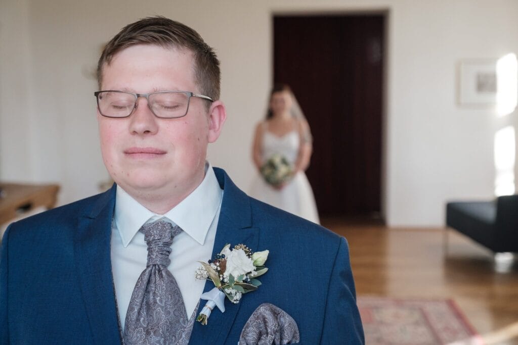 Hochzeitskrone Ein Mann im Anzug mit geschlossenen Augen steht im Vordergrund und fängt einen dieser bezaubernden Momente ein, während im Hintergrund eine Frau im Brautkleid einen Blumenstrauß hält. Diese Szene verkörpert wunderbar natürliche Hochzeitsfotos und unterstreicht exquisite Hochzeitsfotografie in Niederösterreich.