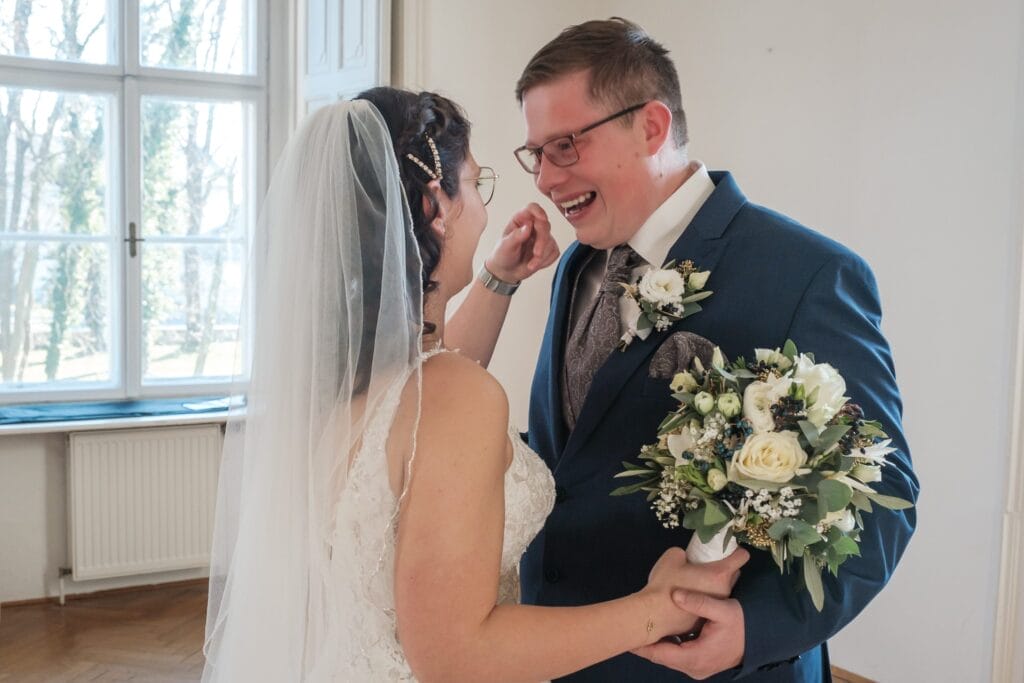 Hochzeitskrone In einem hellen Raum tauschen Braut und Bräutigam bezaubernde Lächeln aus und fangen bezaubernde Momente ein. Der Brautstrauß ergänzt Anzug, Krawatte und Ansteckblume des Bräutigams perfekt – ein Beweis für natürliche Hochzeitsfotos, die ihre Freude an atemberaubender Hochzeitsfotografie in Niederösterreich widerspiegeln.