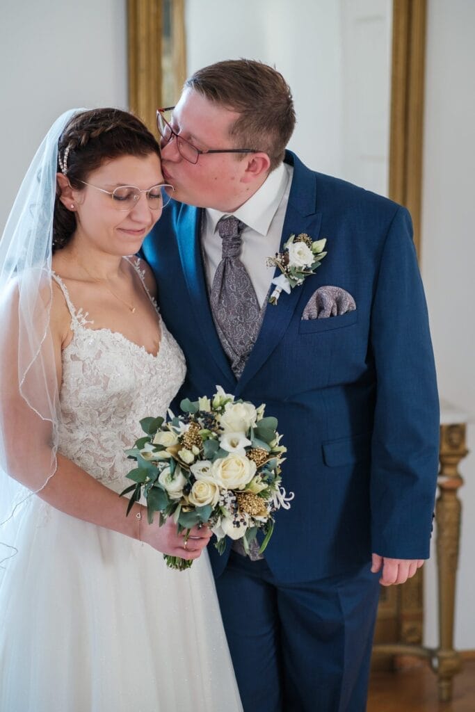 Hochzeitskrone Eine Braut in einem weißen Kleid und Schleier hält einen Blumenstrauß, während sie neben einem Bräutigam im blauen Anzug steht, der sie auf die Stirn küsst und einen dieser bezaubernden Momente festhält, die natürliche Hochzeitsfotos ausmachen.