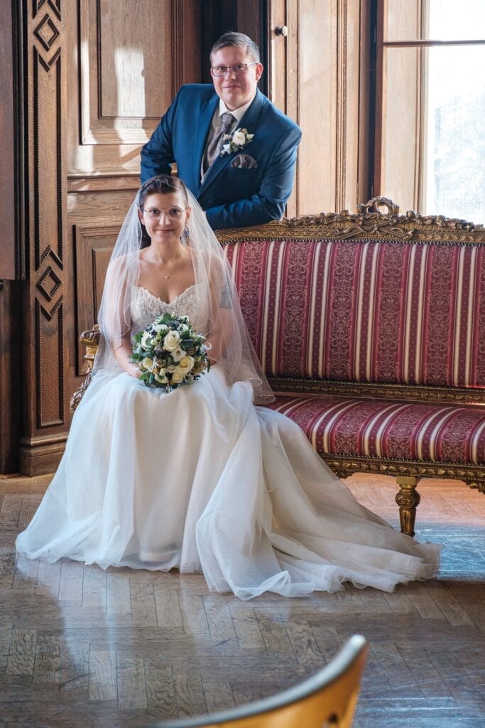 Hochzeitskrone In einem holzgetäfelten Raum sitzt eine Braut im weißen Kleid mit einem Blumenstrauß in der Hand auf einem rot gestreiften Sofa, während der Bräutigam im blauen Anzug hinter ihr herzlich lächelt. Ihre bezaubernde Szene fängt bezaubernde Momente der Hochzeitsfotografie in Niederösterreich ein.