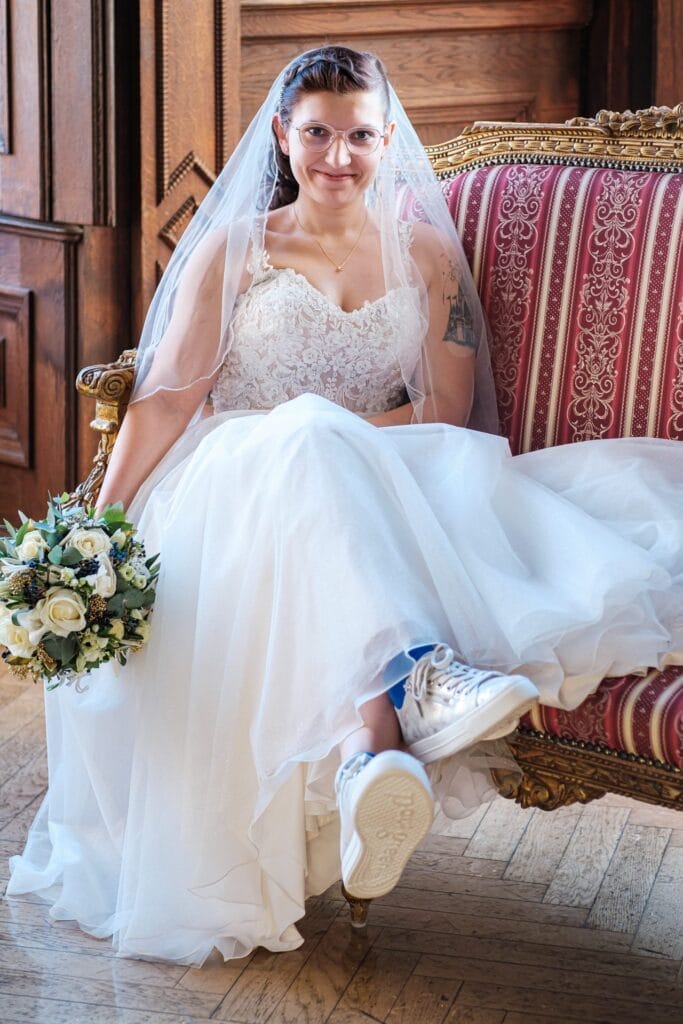 Hochzeitskrone Eine Braut in einem Spitzenbrautkleid und Schleier sitzt anmutig auf einem Vintage-Stuhl, hält einen Blumenstrauß in der Hand und trägt Turnschuhe in einem holzgetäfelten Raum. Halten Sie diese bezaubernden Momente mit natürlichen Hochzeitsfotos fest, die den Charme der Hochzeitsfotografie in Niederösterreich widerspiegeln.