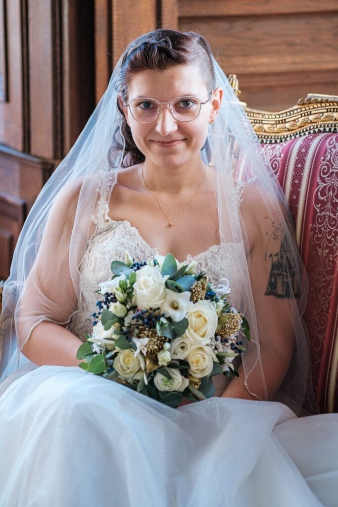 Hochzeitskrone Die Braut sitzt mit Brille und Spitzenschleier da und hält einen Strauß weißer und blauer Blumen in der Hand. Sie fängt auf einem verzierten Stuhl vor einem hölzernen Hintergrund sitzend anmutig bezaubernde Momente ein – ein exquisites Beispiel für Hochzeitsfotografie in Niederösterreich.