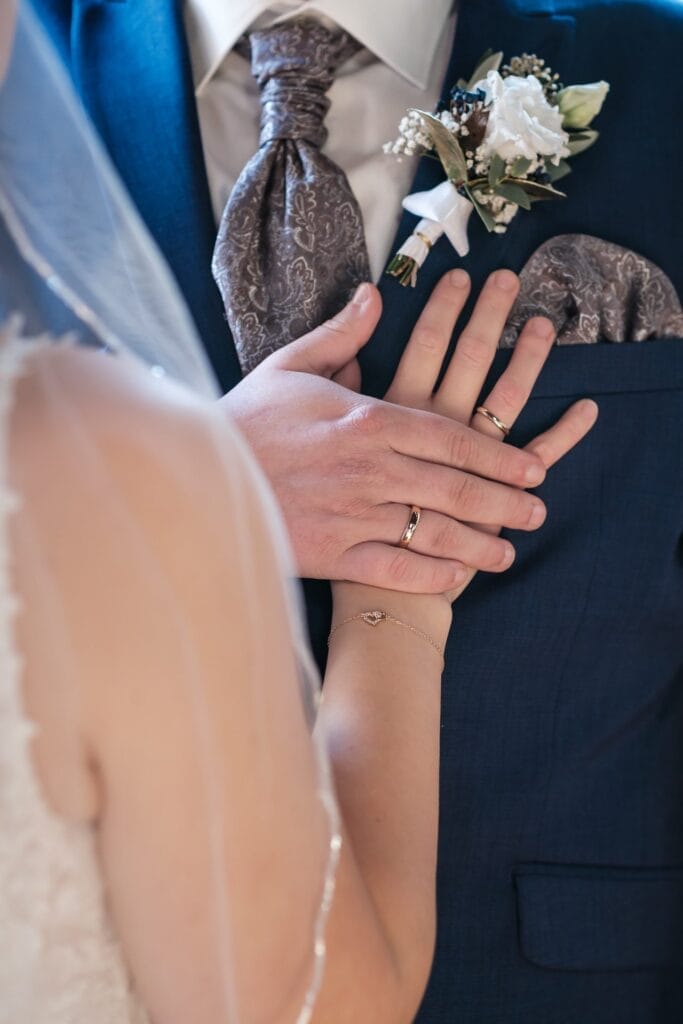 Hochzeitskrone Nahaufnahme der Hände eines Paares mit Eheringen, die bezaubernde Momente festhält. Der Bräutigam, in einem blauen Anzug mit einer Blumenboutonniere, berührt sanft die Hand der Braut, während sie in ihrem weißen Kleid und Schleier glänzt – perfekt für natürliche Hochzeitsfotos.