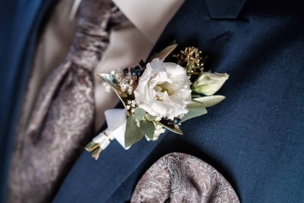 Hochzeitskrone Nahaufnahme einer weißen Blumenboutonniere, die an einer blauen Anzugjacke befestigt ist und bezaubernde Momente einfängt. Auch eine Paisley-gemusterte Krawatte und ein Einstecktuch sind zu sehen, perfekt für natürliche Hochzeitsfotos oder exquisite Hochzeitsfotografie in Niederösterreich.