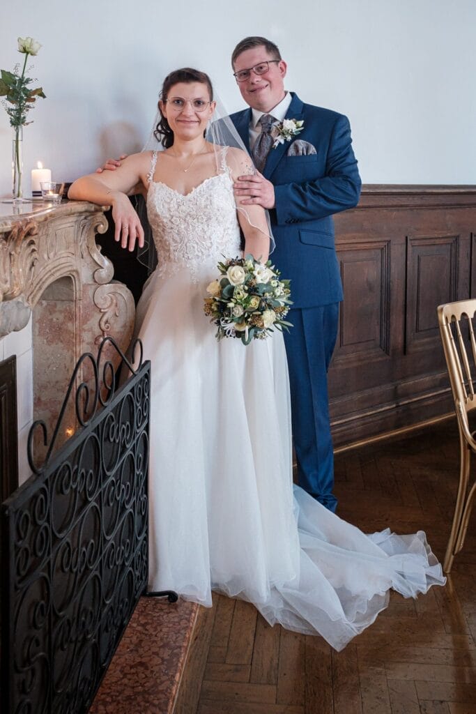 Hochzeitskrone Ein Paar im Hochzeitskleid posiert drinnen vor einem Kamin und fängt bezaubernde Momente ein. Die Braut hält einen Blumenstrauß und der Bräutigam hat ein Glas in der Hand. Beide lächeln sanft und schaffen natürliche Hochzeitsfotos, die pure Freude und Liebe widerspiegeln.