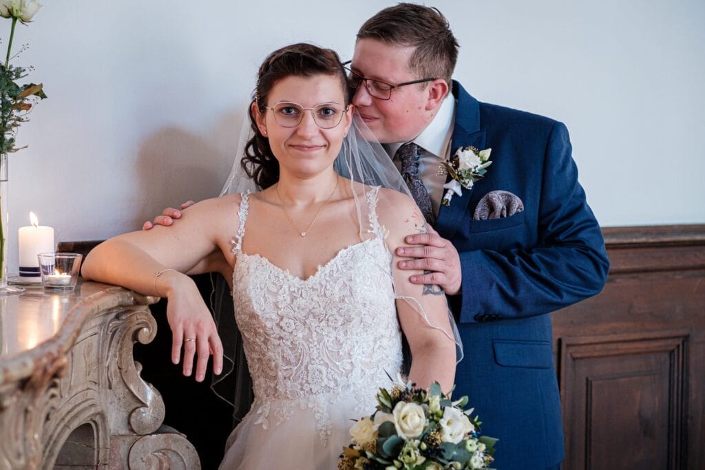 Hochzeitskrone Die Braut im Spitzenkleid hält einen Blumenstrauß und steht neben dem Bräutigam im blauen Anzug. Er berührt ihre Schulter und lehnt sich zu ihr hinüber. Sie sind drinnen, mit einer brennenden Kerze auf einer Marmoroberfläche, und halten bezaubernde Momente ihres besonderen Tages in natürlichen Hochzeitsfotos fest.