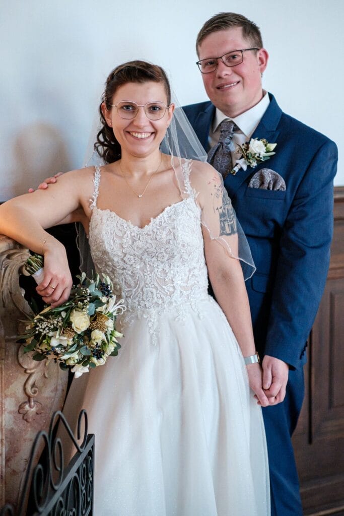 Hochzeitskrone Die Braut in einem weißen Spitzenhochzeitskleid hält einen Blumenstrauß in der Hand, während ihr Bräutigam in einem blauen Anzug steht. Beide lächeln strahlend. Es ist einer dieser bezaubernden Momente, die durch natürliche Hochzeitsfotos festgehalten werden und ihre Freude perfekt zur Geltung bringen.