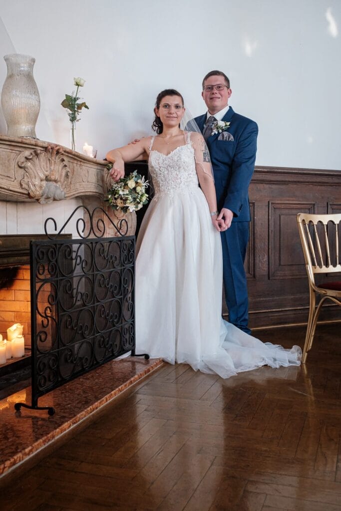 Hochzeitskrone Braut und Bräutigam stehen an einem dekorierten Kamin und fangen bezaubernde Momente ein. Die Braut trägt ein weißes Kleid und hält einen Blumenstrauß, während der Bräutigam einen marineblauen Anzug mit einer Boutonniere trägt. Kerzen flackern sanft auf und neben dem Kaminsims und verkörpern natürliche Hochzeitsfotos.