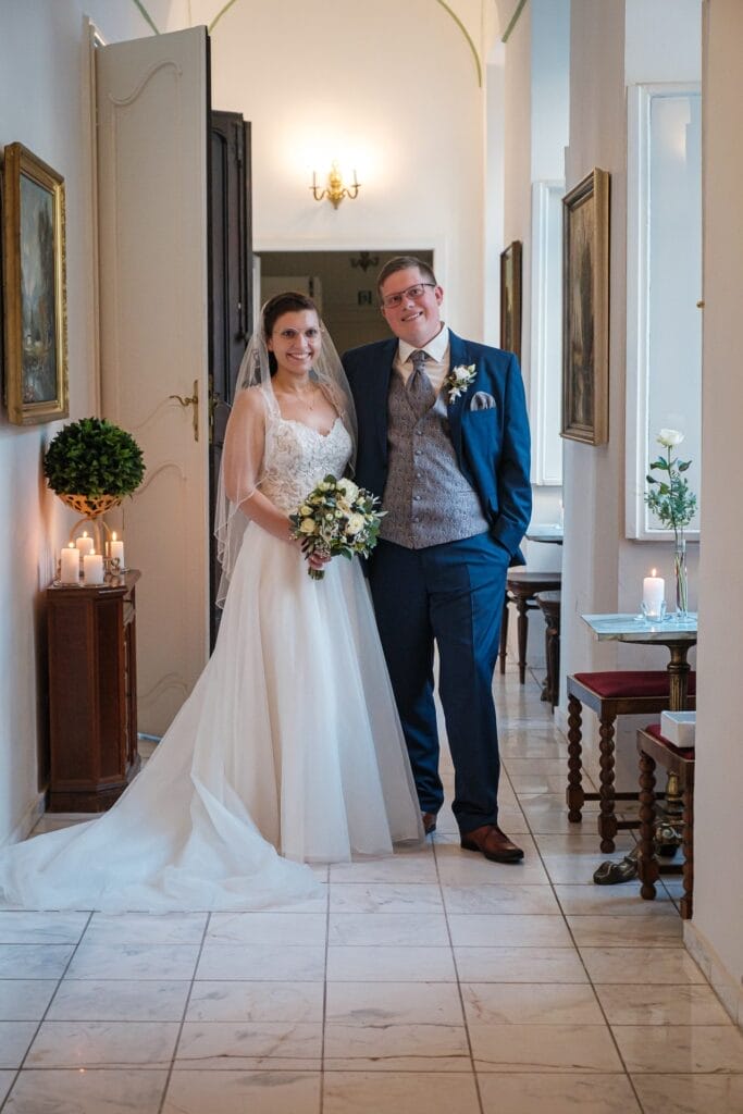 Hochzeitskrone Braut und Bräutigam stehen gemeinsam in einem Flur; die Braut hält einen Blumenstrauß, während der Bräutigam einen blauen Anzug trägt. Kerzen und Topfpflanzen schmücken den Hintergrund und fangen bezaubernde Momente der Hochzeitsfotografie in Niederösterreich ein.