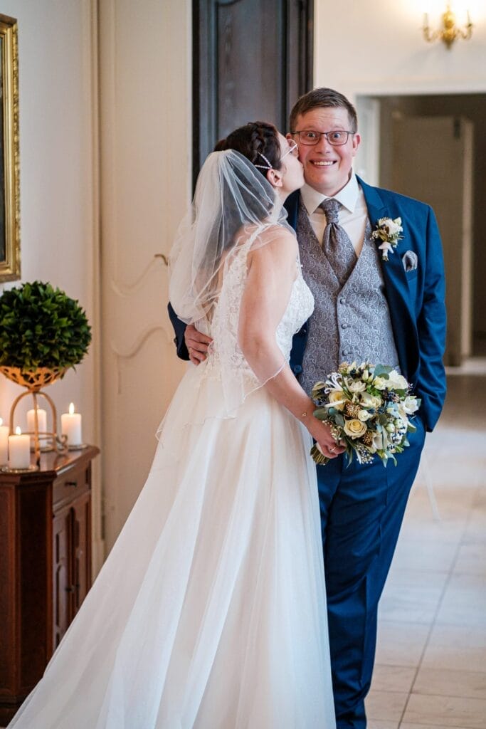 Hochzeitskrone Die Braut im weißen Kleid küsst den Bräutigam im blauen Anzug im Innenbereich, hält einen Blumenstrauß in der Hand und fängt bezaubernde Momente ein, die die Essenz natürlicher Hochzeitsfotos widerspiegeln.