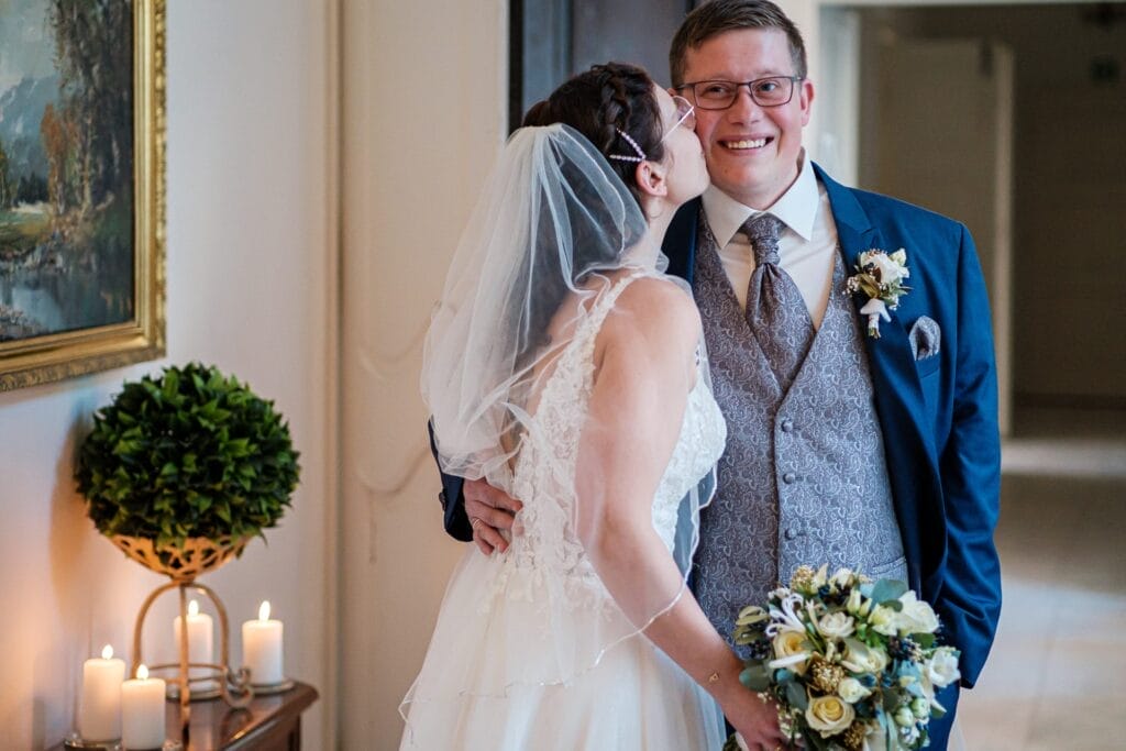 Hochzeitskrone Eine Braut im weißen Kleid küsst den Bräutigam auf die Wange und fängt bezaubernde Momente ein. Der Bräutigam, in einem blauen Anzug, hält einen Blumenstrauß und ist von brennenden Kerzen und einer Pflanze umgeben. Diese natürlichen Hochzeitsfotos verkörpern wunderschön die Magie ihres besonderen Tages im Haus.