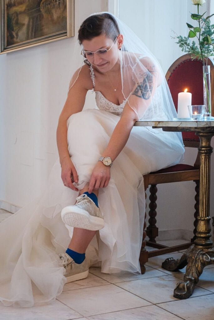 Hochzeitskrone Eine Braut im Brautkleid und Schleier sitzt auf einem Stuhl und bindet die Schnürsenkel ihrer weißen Turnschuhe und blauen Socken. Neben ihr sorgen eine Kerze und Blumen für einen besonderen Charme – bezaubernde Momente in natürlichen Hochzeitsfotos festhalten.