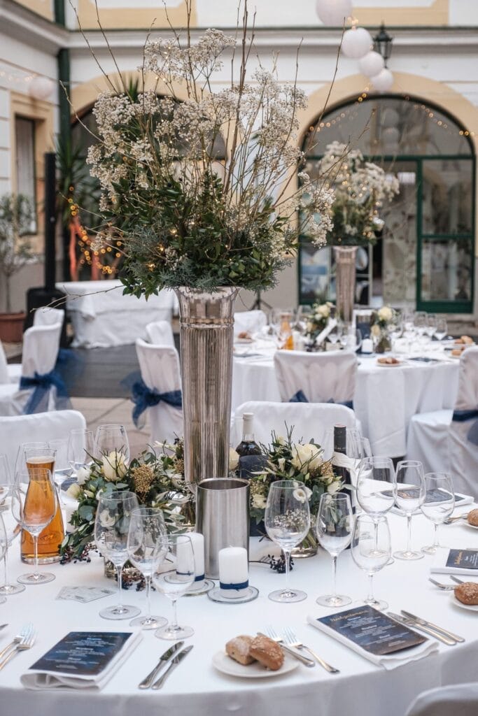 Hochzeitskrone Elegant gedeckter Tisch für den Hochzeitsempfang mit hohem Blumengesteck, weißen Tischdecken, ordentlich arrangierten Gedecken, Weingläsern und Brötchen in einem Innenhof mit dekorativer Beleuchtung – perfekt, um bezaubernde Momente zu schaffen, festgehalten in natürlichen Hochzeitsfotos.