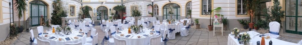 Hochzeitskrone Panoramablick auf einen elegant dekorierten Essbereich im Freien mit stilvoll gedeckten runden Tischen, die bezaubernde Momente einfangen. Weiße Tischdecken, blaue Schärpen und Geschirr sind von Pflanzen und Bogenfenstern umgeben – die perfekte Kulisse für natürliche Hochzeitsfotos.