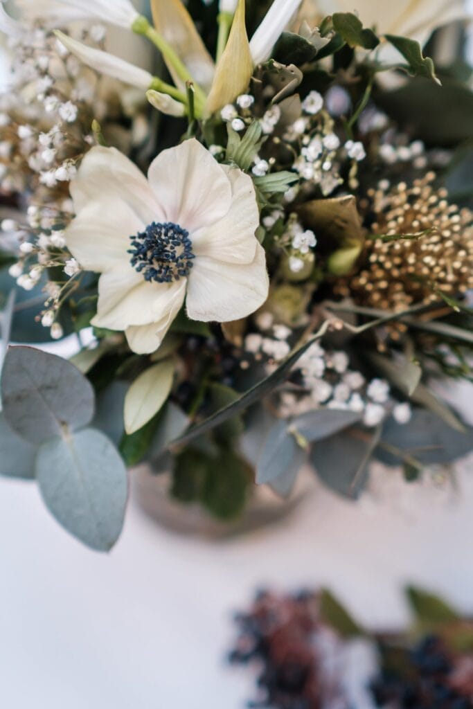 Hochzeitskrone Eine Nahaufnahme eines Blumenarrangements fängt bezaubernde Momente mit einer weißen Blume mit dunkler Mitte ein, umgeben von Grün und kleinen weißen Blüten, perfekt für natürliche Hochzeitsfotos in Niederösterreich.