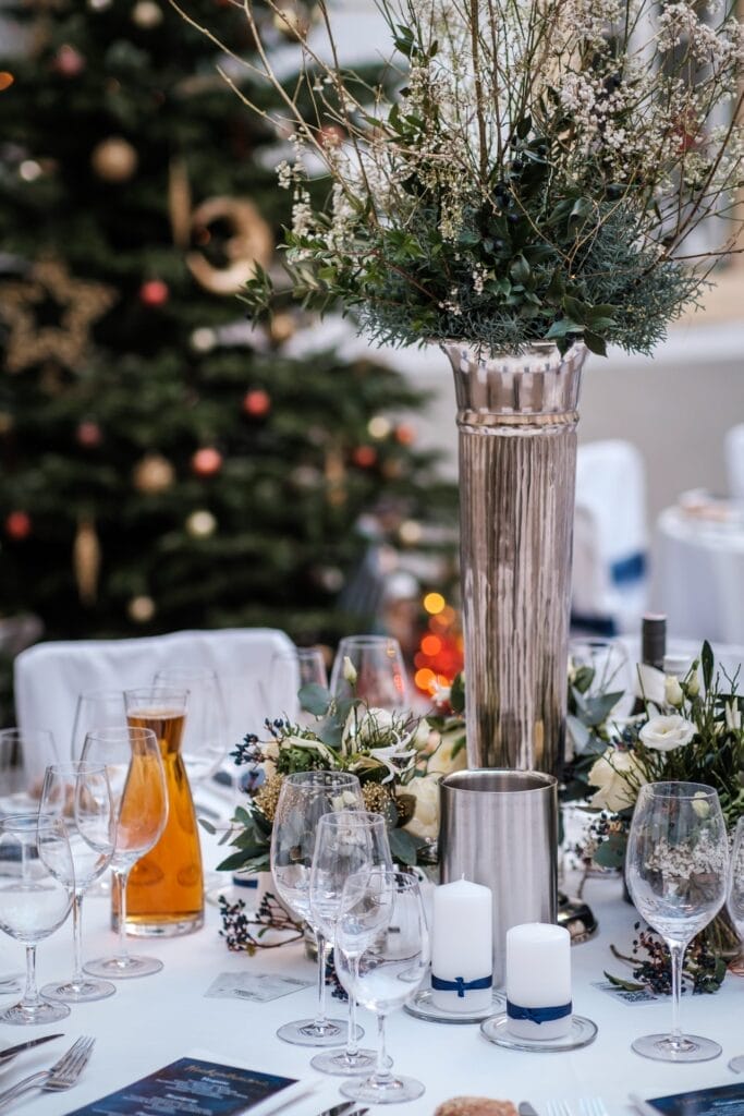 Hochzeitskrone Eine elegante Tischdekoration mit Weingläsern, einem großen Blumengesteck, Kerzen und einer Karaffe auf einer weißen Tischdecke setzt die Szene. Ein geschmückter Weihnachtsbaum verleiht dem Hintergrund Charme, perfekt für bezaubernde Momente in der Hochzeitsfotografie in Niederösterreich.