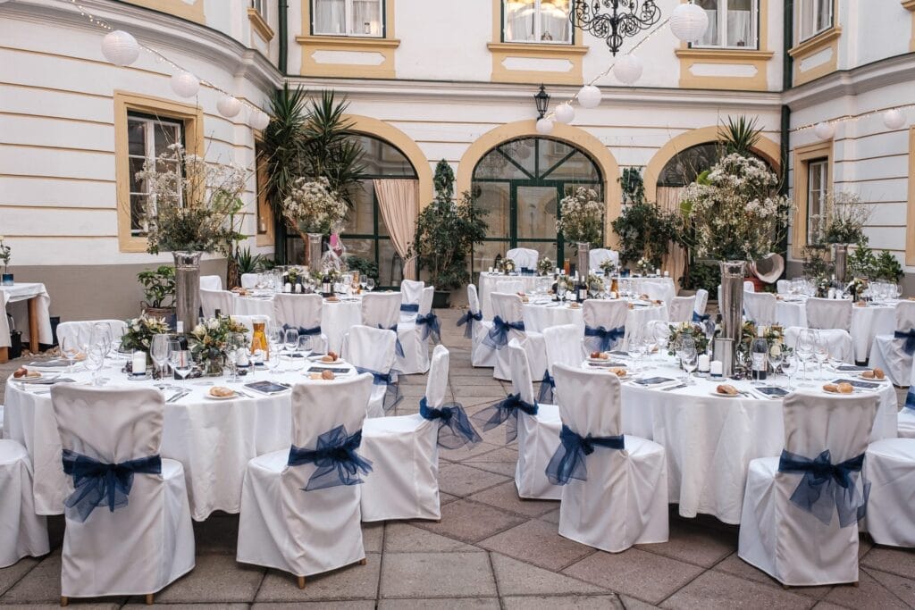 Hochzeitskrone Eine malerische Hochzeitsfeier im Freien besteht aus runden Tischen und weißen Stühlen, die mit blauen Schleifen geschmückt sind, in einem bezaubernden Innenhof. Weiße Tischdecken, elegante Tafelaufsätze, Teller und Gläser vervollständigen diese bezaubernde Szene – die perfekte Kulisse, um bezaubernde Momente festzuhalten.