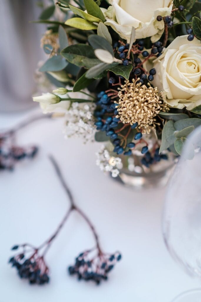 Hochzeitskrone Ein Strauß weißer Rosen, goldfarbener Blüten, grüner Blätter und blauer Beeren ziert eine silberne Vase auf dem Tisch. Zwei Zweige blauer Beeren liegen daneben und fangen bezaubernde Momente ein, die an natürliche Hochzeitsfotos erinnern und für immer in Erinnerung bleiben.