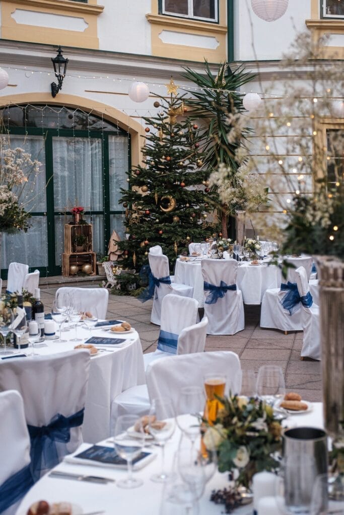 Hochzeitskrone Ein bezauberndes Outdoor-Event mit runden Tischen, die mit weißen Tüchern und blauen Schleifen bedeckt und mit Blumenarrangements geschmückt sind. Ein majestätischer Weihnachtsbaum ziert den Hintergrund und schafft bezaubernde Momente, ideal für natürliche Hochzeitsfotos.