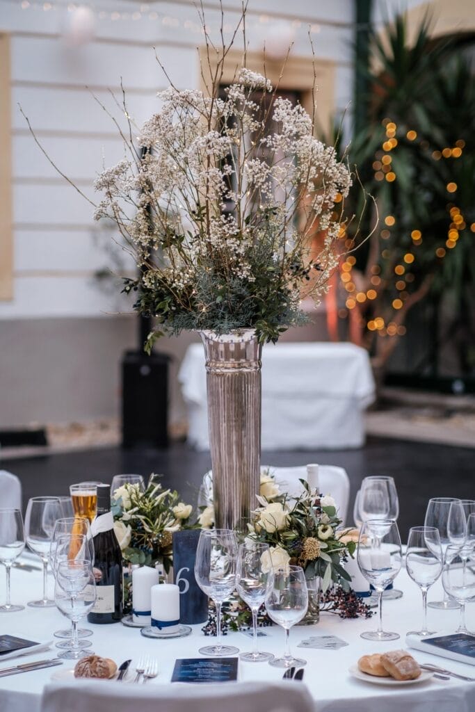 Hochzeitskrone Elegant dekorierter Tisch mit einem hohen Blumengesteck, Weingläsern, Kerzen, Brötchen und einer Flasche Wein auf einer weißen Tischdecke. Die festliche Kulisse fängt mit ihren zauberhaften Lichterketten bezaubernde Momente ein.