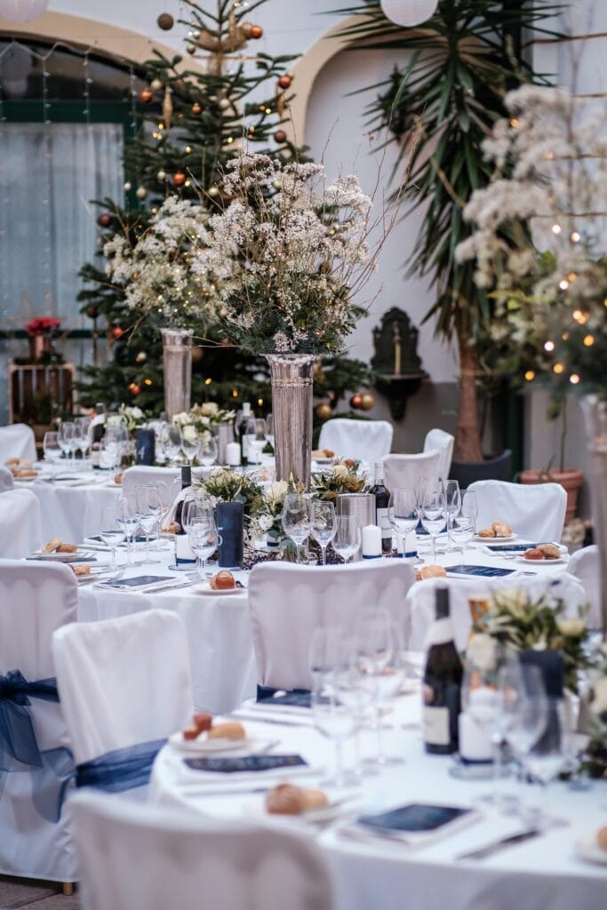 Hochzeitskrone Elegantes Event-Setup mit runden Tischen, weißen Tischdecken, Weingläsern, Brötchen und hohen Blumengestecken. Die Dekoration umfasst Grünpflanzen und kleine Bäume mit Lichterketten im Hintergrund – perfekt für bezaubernde Momente, festgehalten von Hochzeitsfotografie in Niederösterreich.